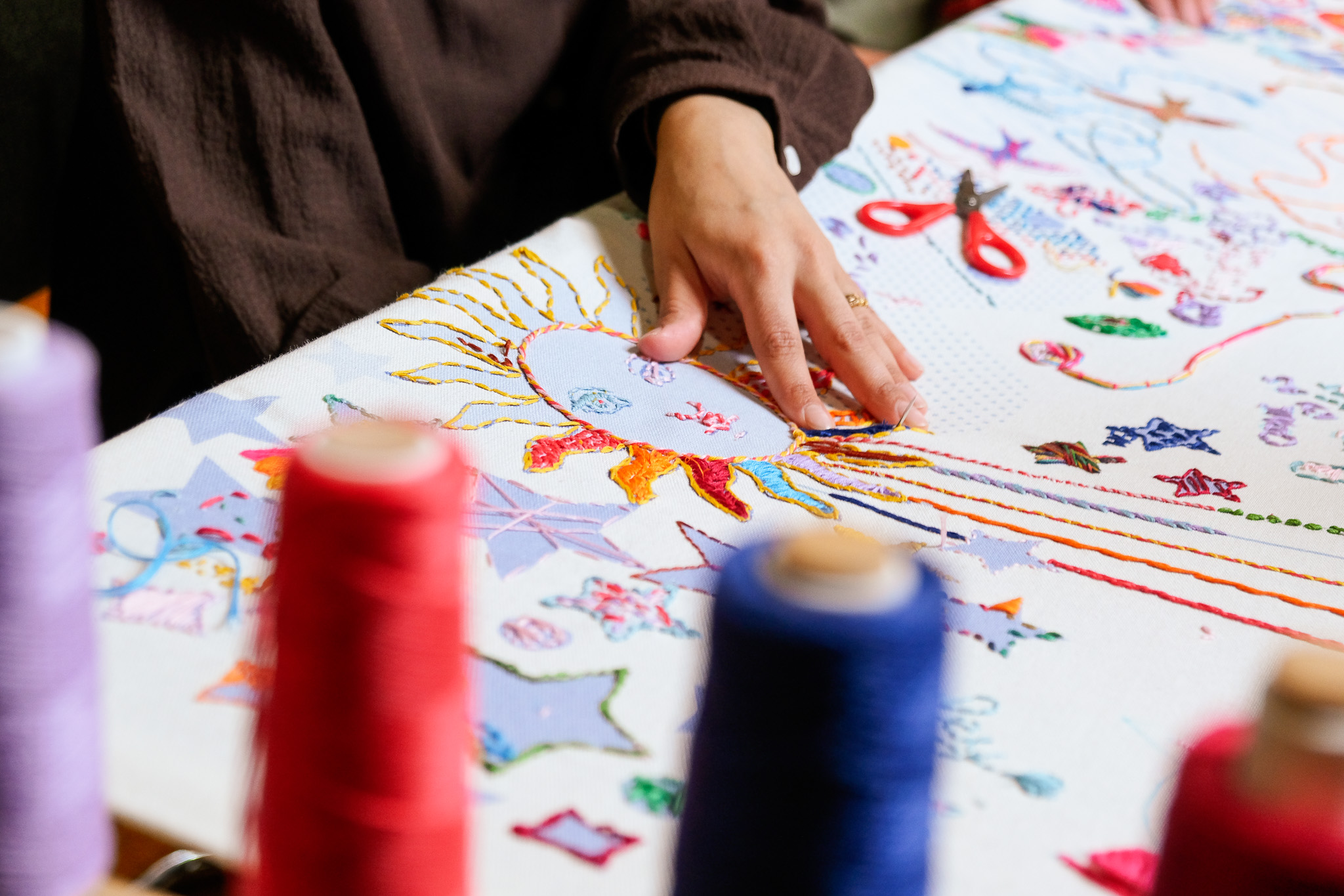 Hands stitching a colourful sun which has a face. In the foreground are spools of colourful thread and surrounding the sun are semi-finished embroidered patterns and stars.