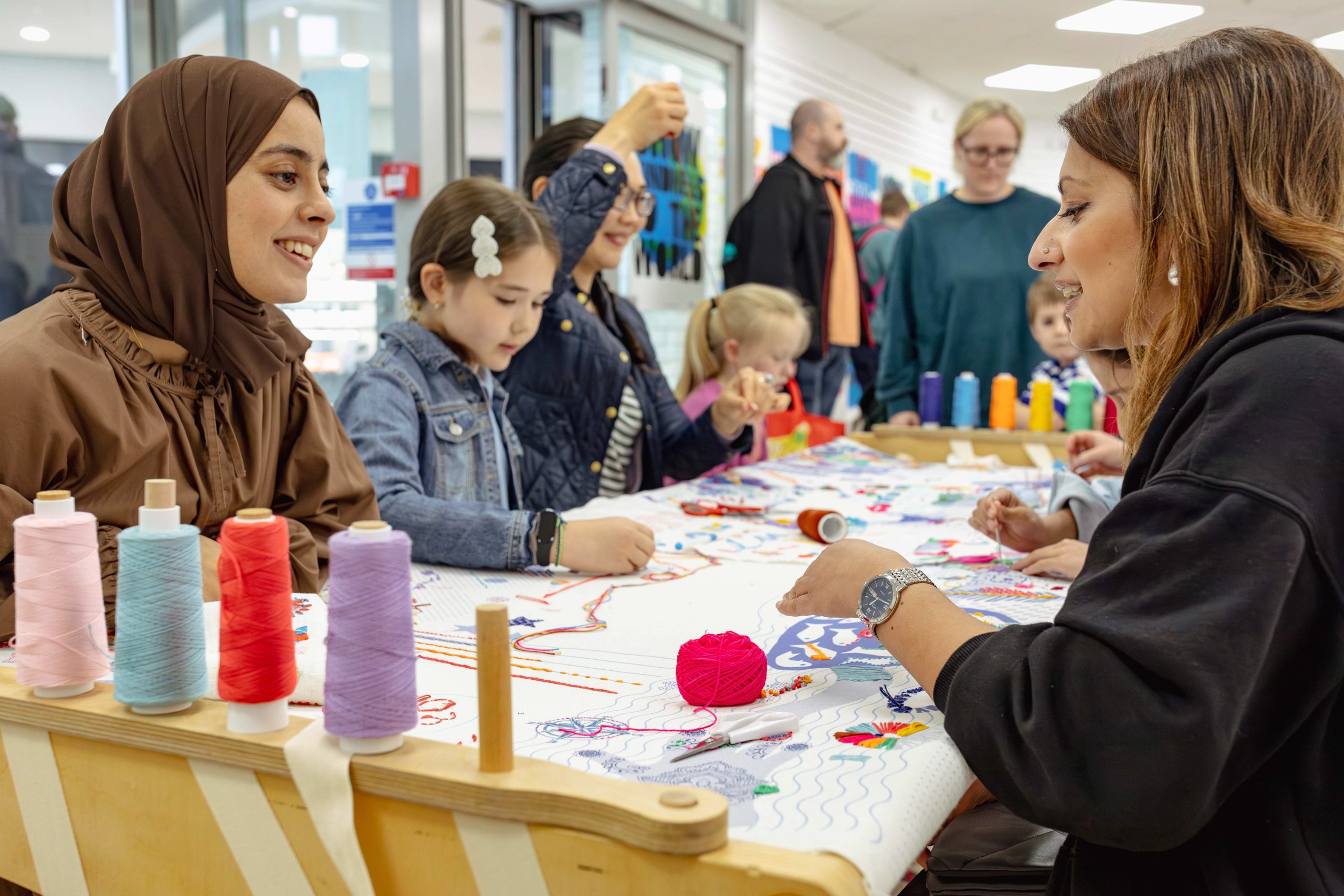 Sana Maulvi is wearing brown and is sat on one side of the stitch table, talking to a woman wearing black who is stitching at the table. Children and adults surround them, joining in with stitching.