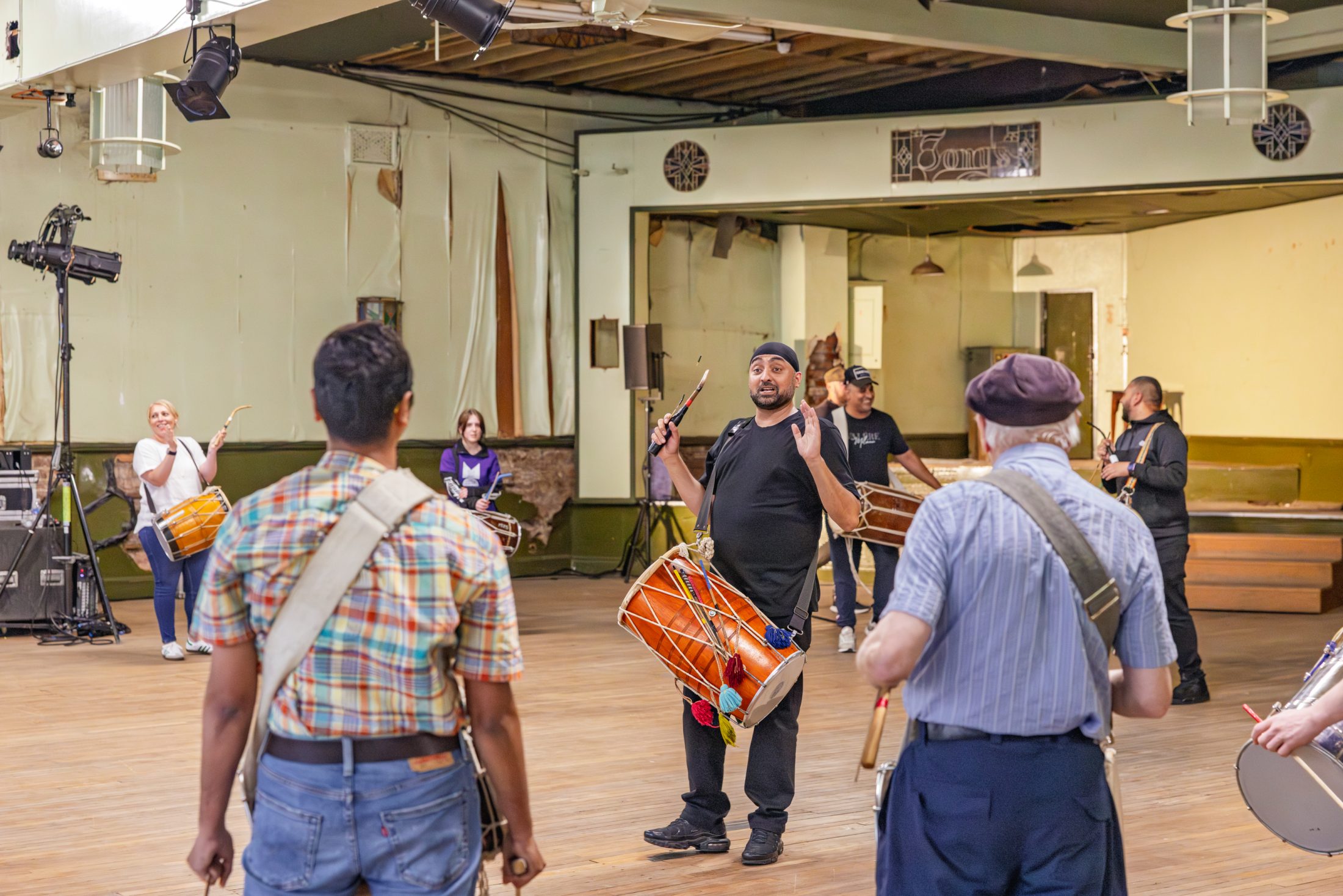 A group of people in a large room practise drumming together, led by an instructor in the centre holding a drum and raising his arm. The participants wear drums strapped across their bodies.