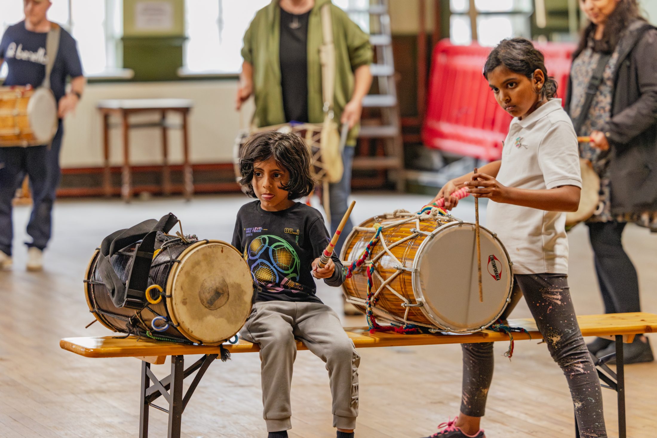 Two children sit on a bench indoors, each with a traditional drum. One child holds a drumstick and plays, whilst the other watches. Several adults with drums stand in the blurred background.