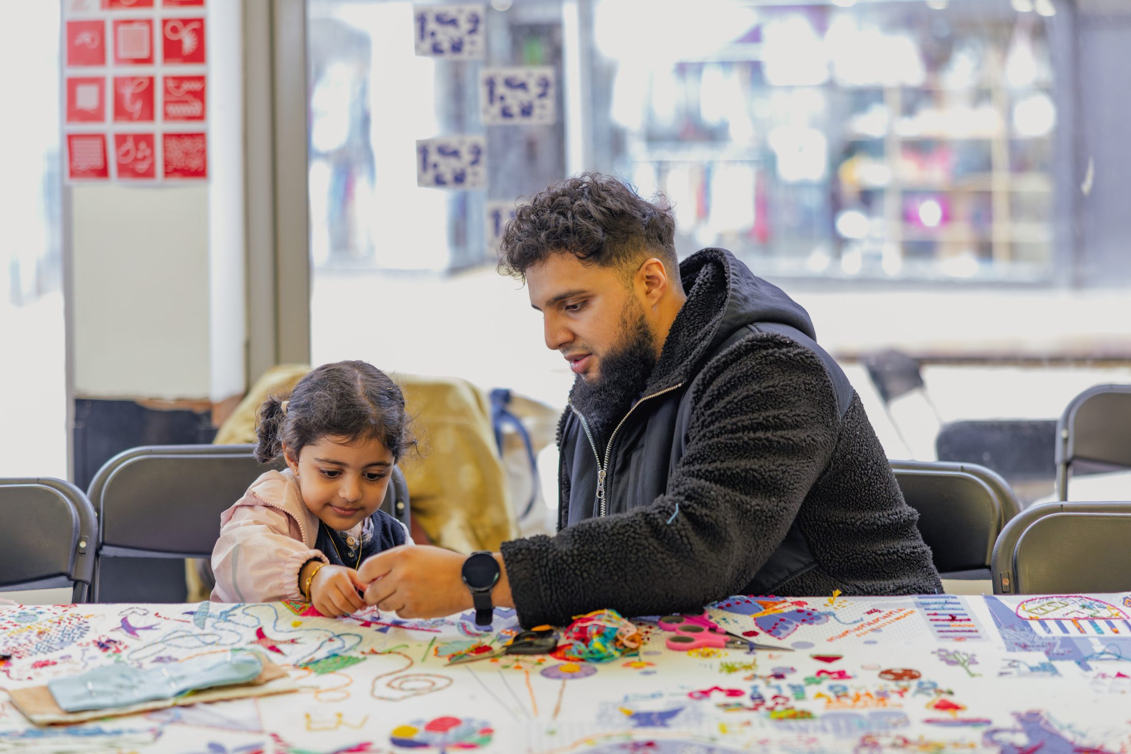 A man and a young girl sit at a table covered with colourful art supplies, working on a craft project together in a bright, indoor creative space.