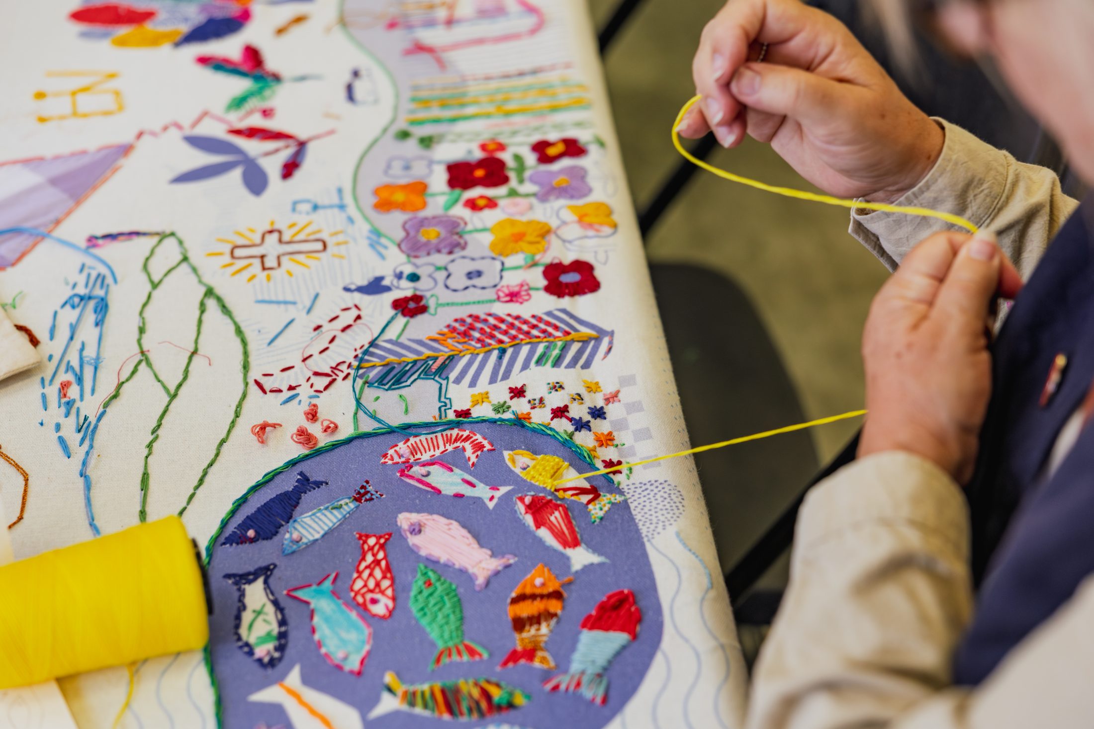 A person hand-stitching colourful embroidery on fabric featuring various patterns, flowers, fish, and thread designs, with a reel of yellow thread nearby on the table.
