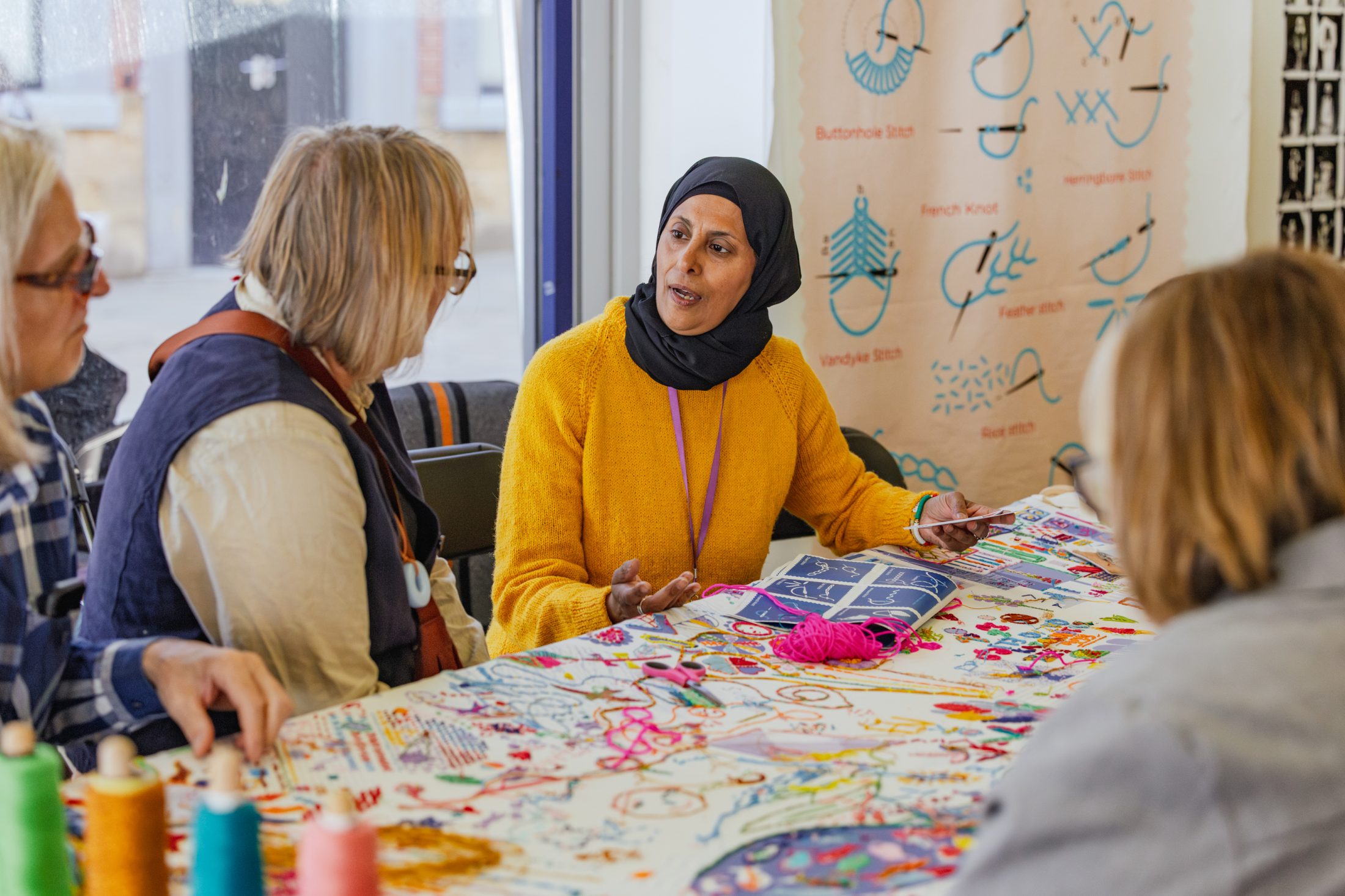 Four women sit around a colourful table with embroidery threads, engaged in conversation. One woman in a yellow jumper and black headscarf is speaking whilst holding a booklet. Embroidery patterns are displayed on the wall.