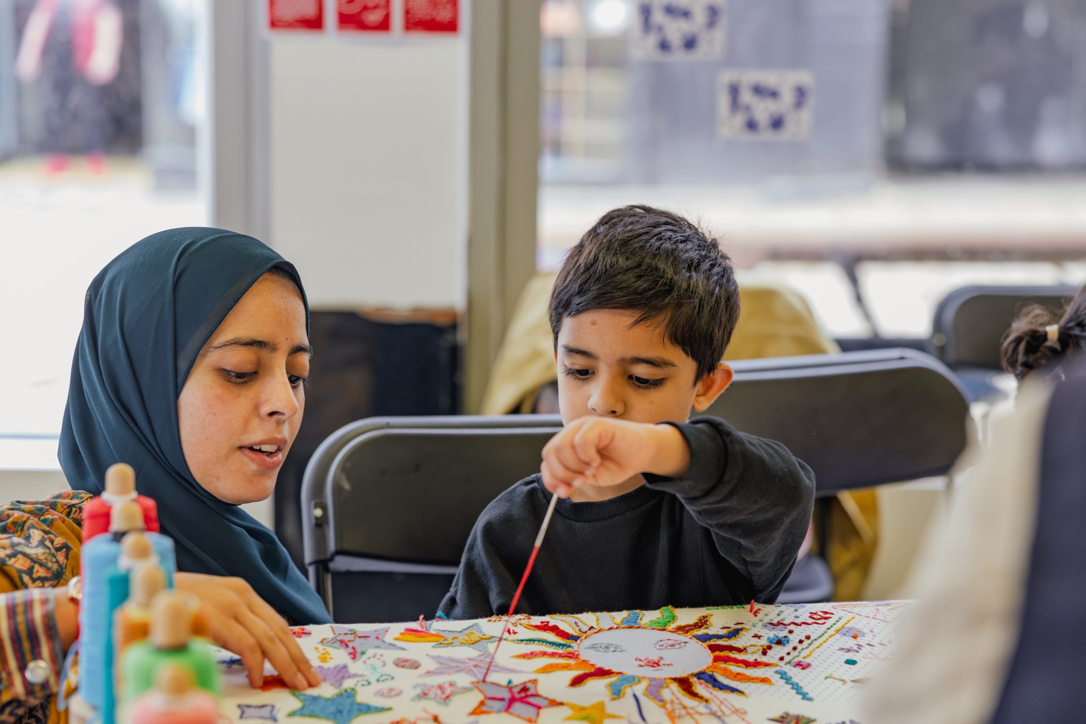 A woman in a hijab and a young boy sit at a table, working together on a colourful craft project with felt-tip pens, paint, and paper in a bright room.