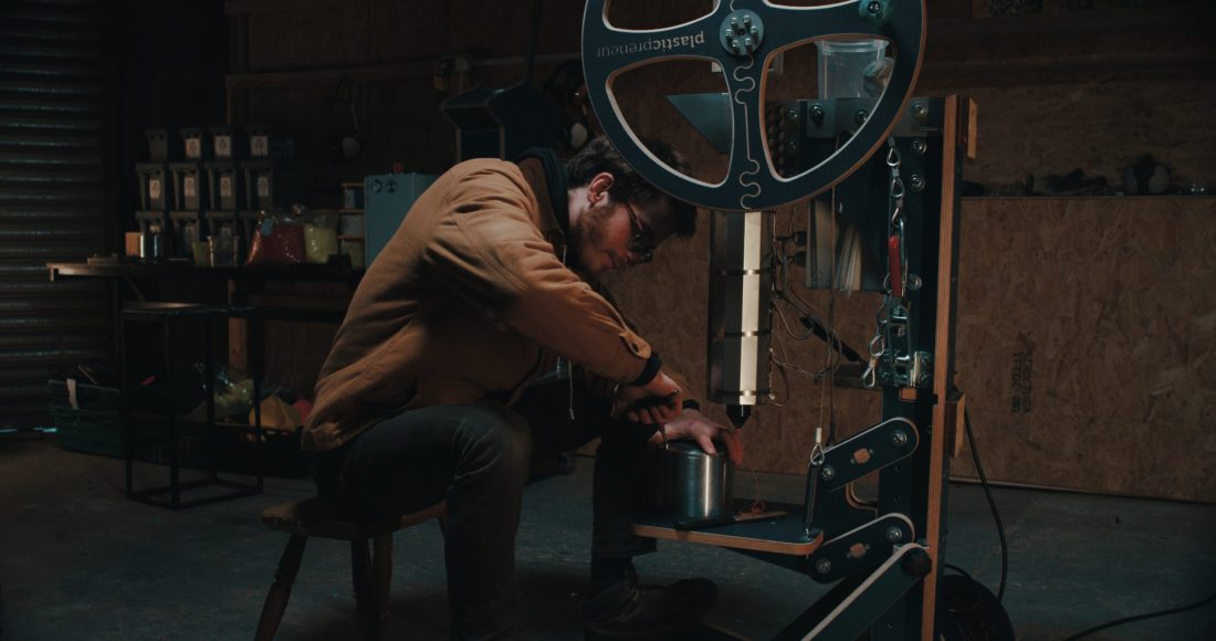 A person wearing glasses and a brown jacket sits on a stool, operating a large mechanical machine in a dimly lit workshop with tools and equipment in the background.
