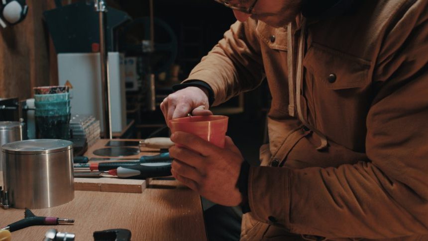 A person wearing glasses and a brown jacket works intently with a small red container at a cluttered wooden workbench, surrounded by various tools and supplies in a workshop setting.