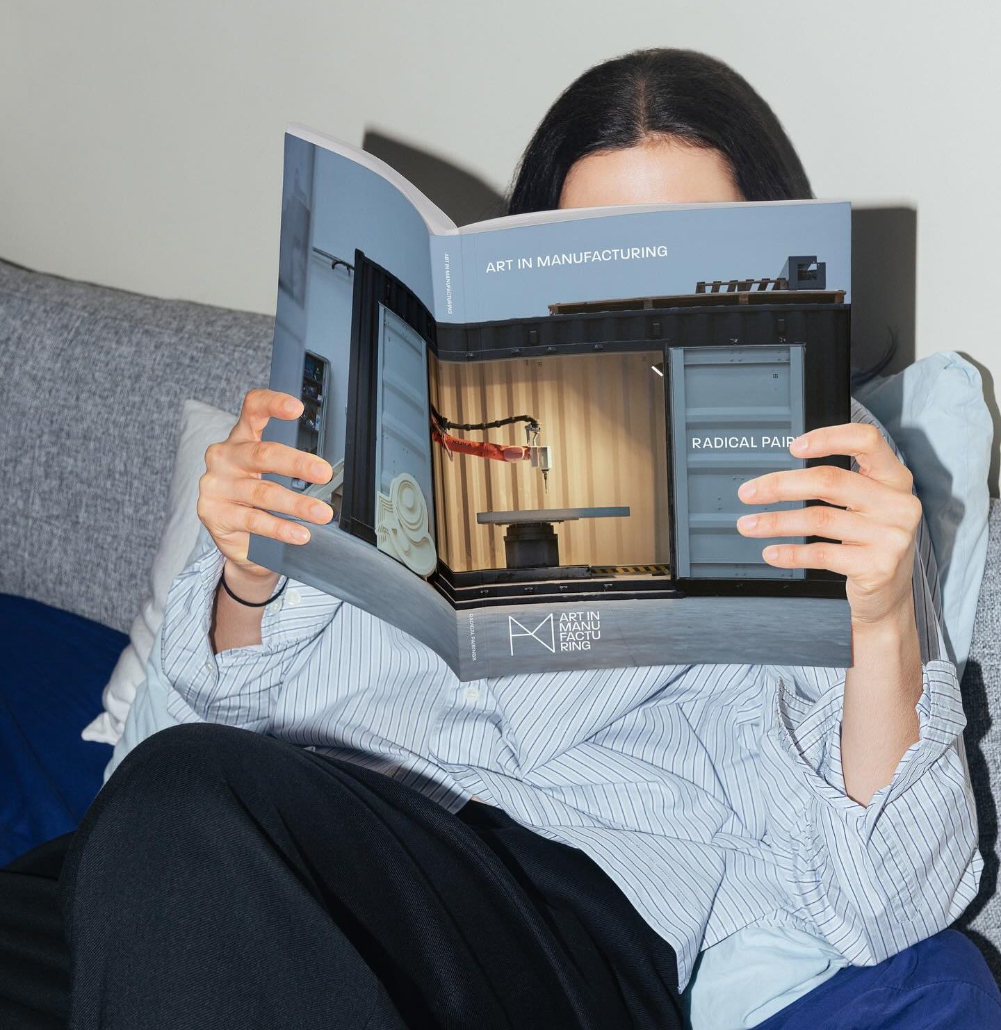 A person with long dark hair sits on a sofa, holding and reading a large magazine titled Art in Manufacturing that covers most of their face. They wear a light blue shirt and black trousers.