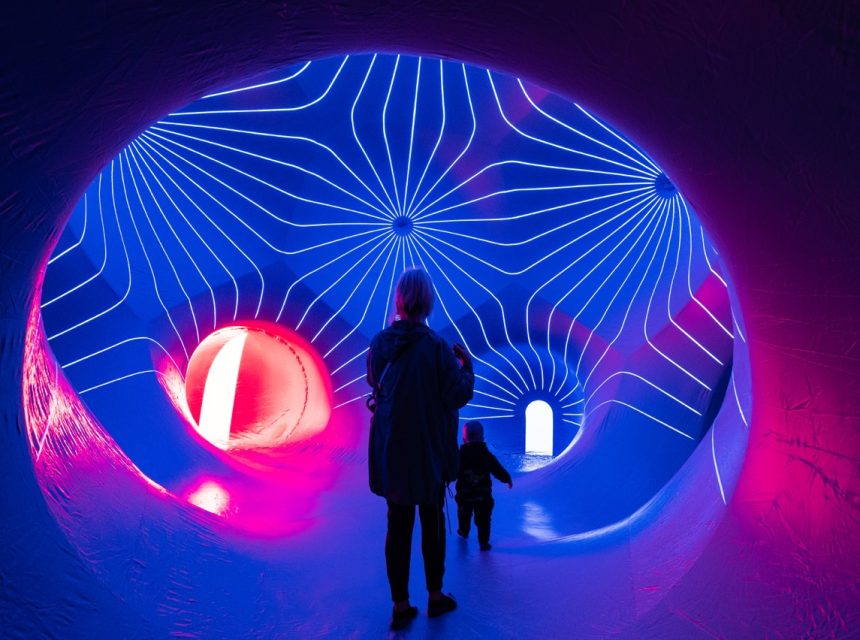A person and a child walk through a glowing tunnel with blue and pink neon lights and curved, radiating lines on the ceiling, creating a futuristic atmosphere.