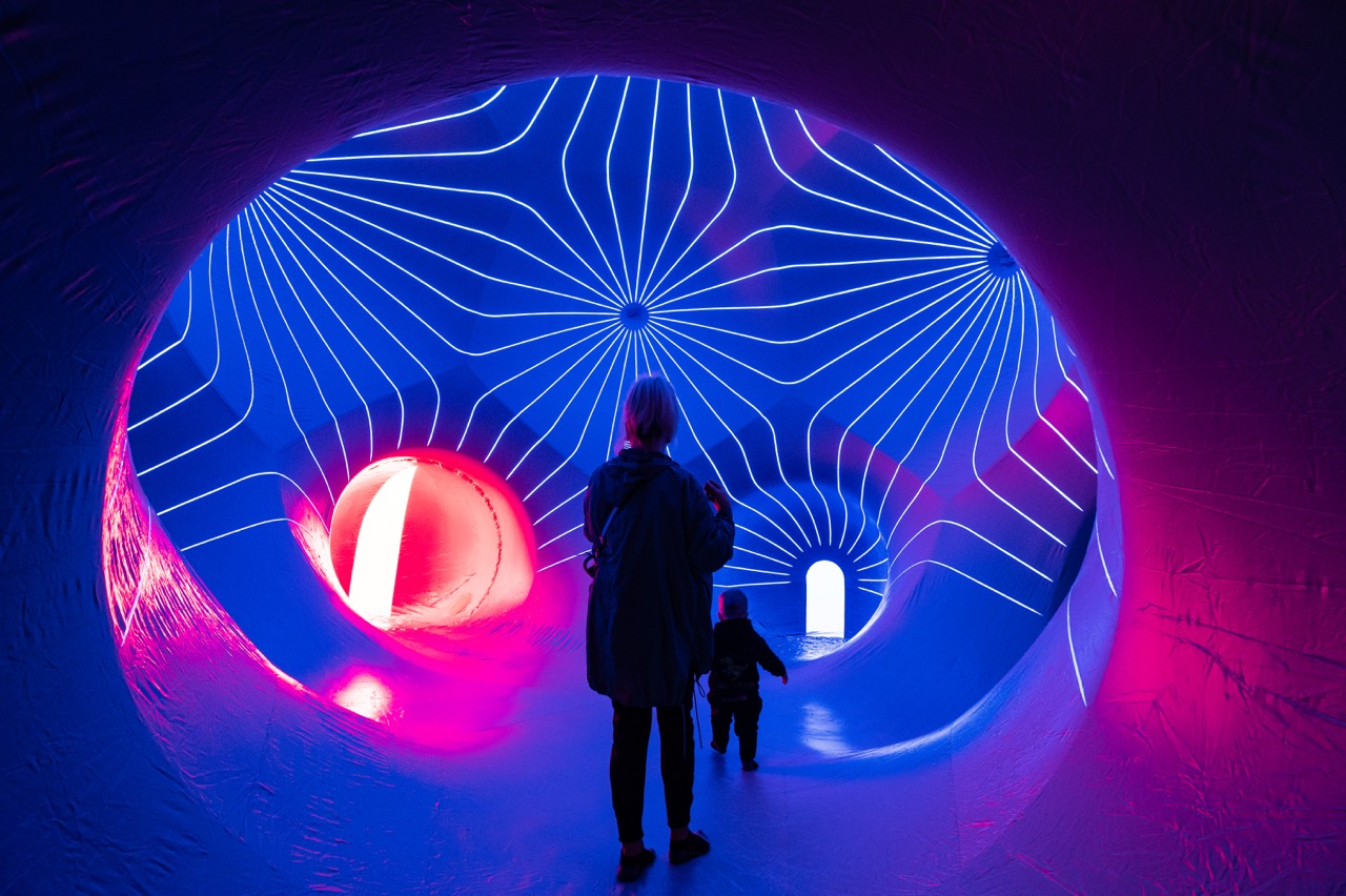 A person and a child walk through a glowing tunnel with blue and pink neon lights and curved, radiating lines on the ceiling, creating a futuristic atmosphere.