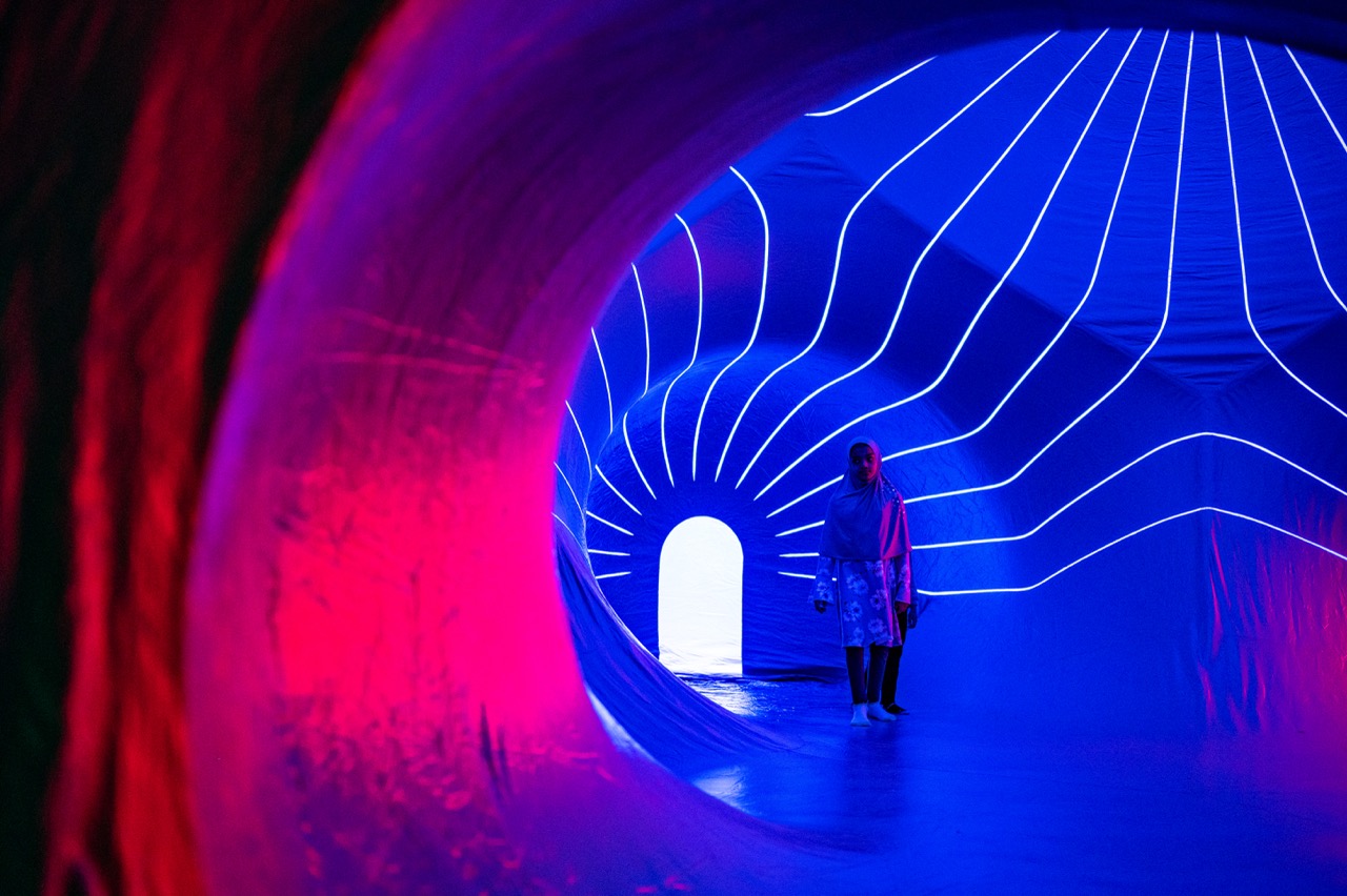 A person stands inside a brightly lit, futuristic tunnel with glowing blue and pink lights emanating from curved walls and radiating white lines surrounding a small arched door.