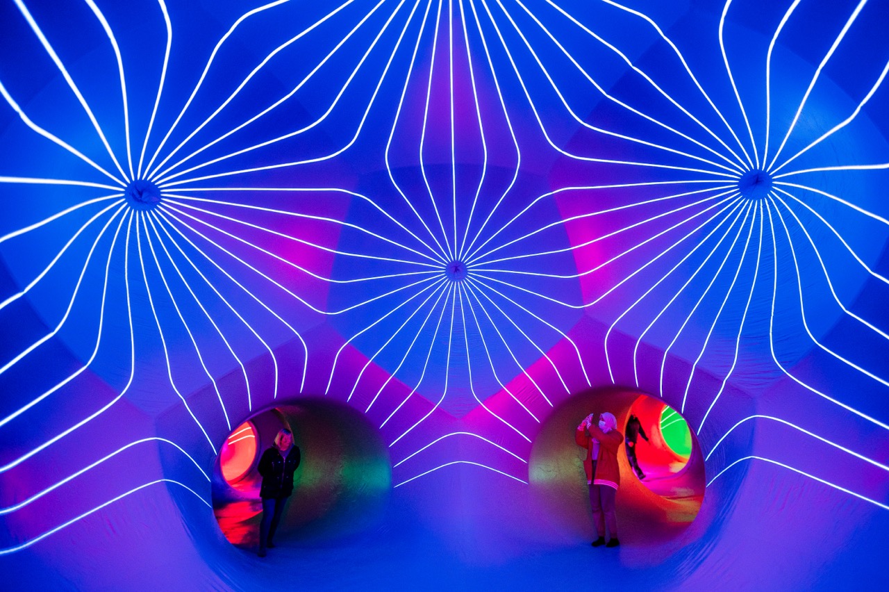 People stand inside a tunnel-like art installation with vibrant blue, pink, and purple lighting. White glowing lines radiate from two central points on the ceiling, creating a mesmerising geometric pattern.