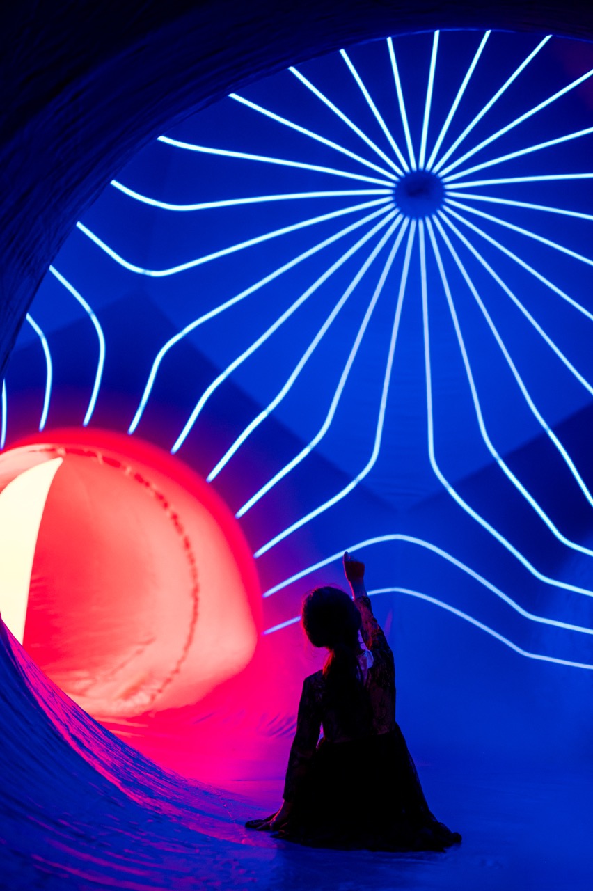 A person sits on the floor inside a tunnel-like space illuminated by vibrant blue and red lights, with glowing white lines radiating from a central point on the ceiling.