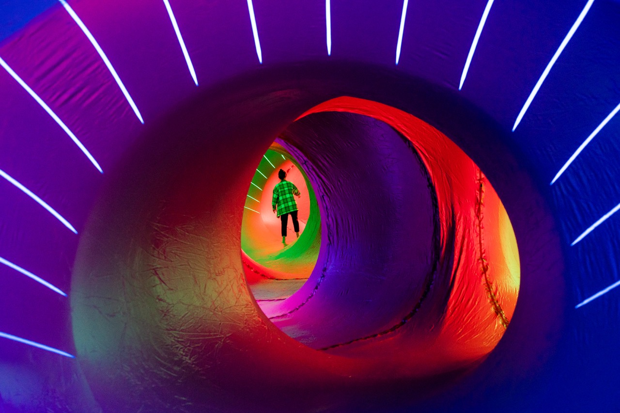 A person wearing a green checked shirt walks inside a colourful, tunnel-like structure illuminated with vibrant purple, red, yellow, and blue lights, creating a surreal, futuristic atmosphere.