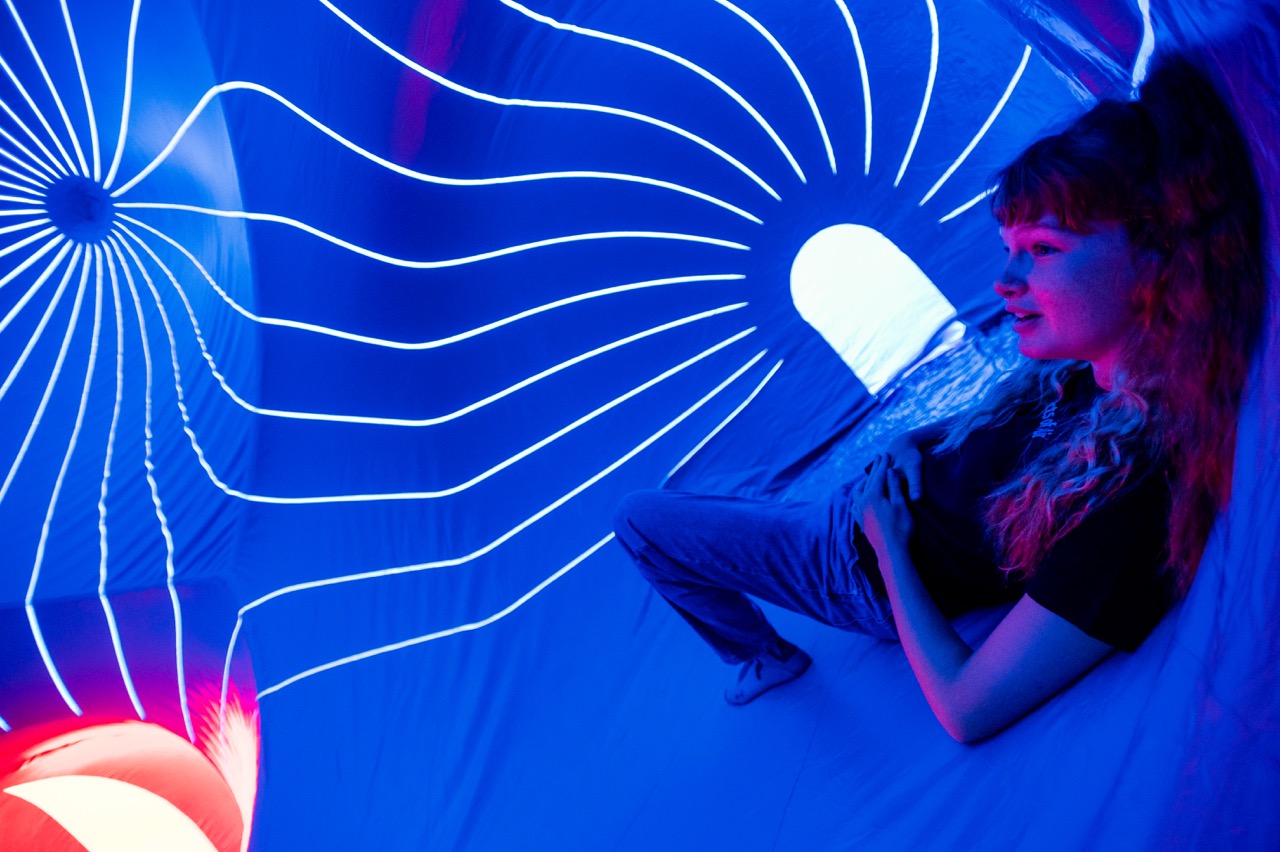 A person relaxes inside a blue-lit, inflatable space with glowing white lines radiating from a central point and an arched window letting in light. The atmosphere is futuristic and immersive.