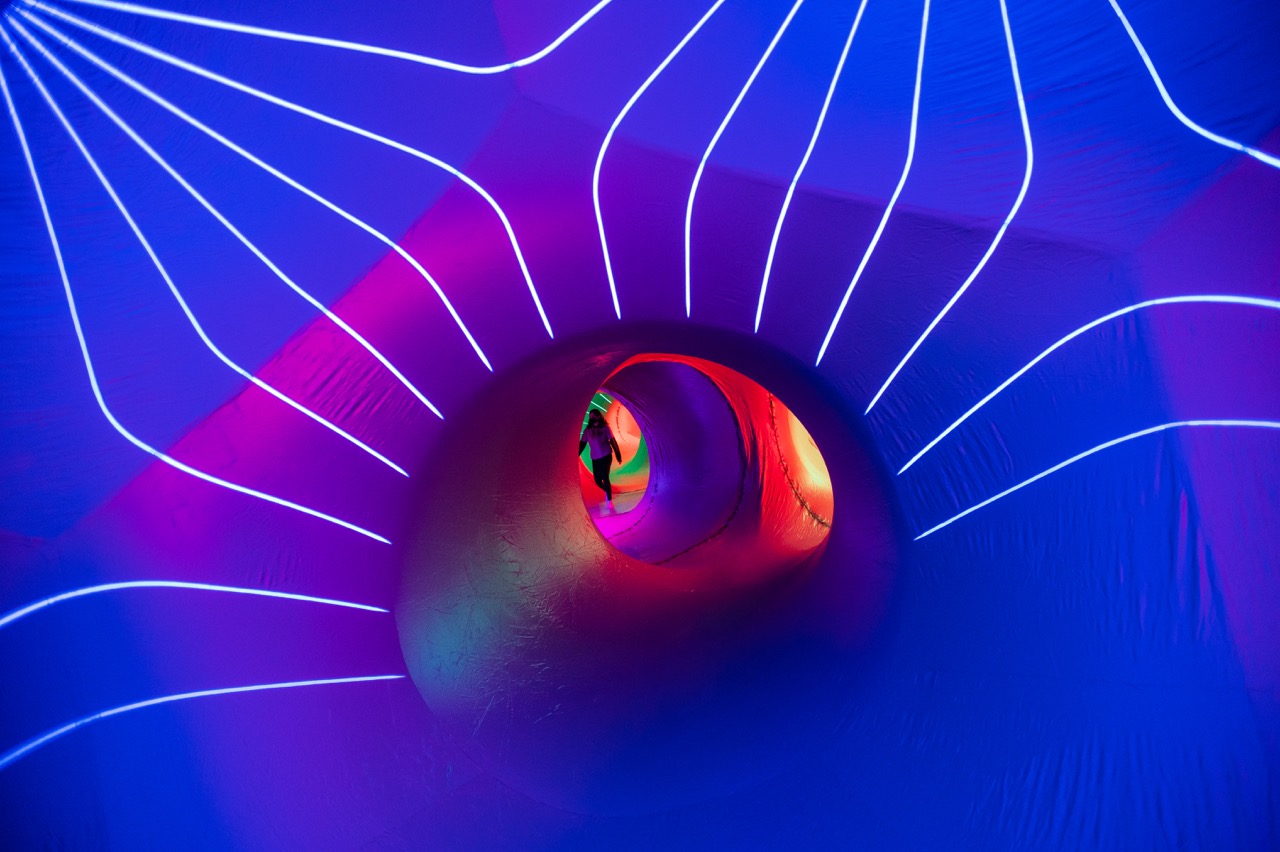 A person stands in the distance inside a tunnel-like structure illuminated with vibrant purple, blue, and pink lights, with glowing lines radiating outward from a central circular opening.