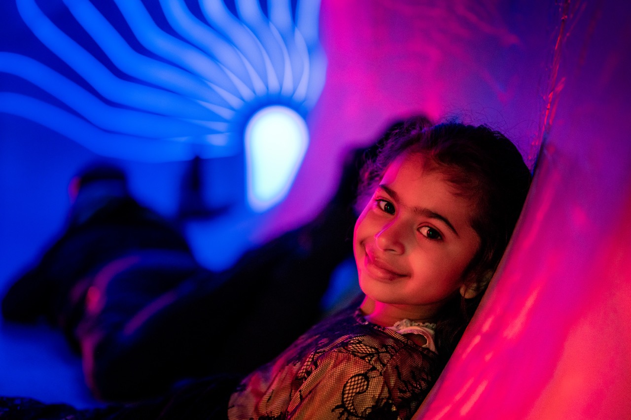 A young girl smiles while reclining inside a tunnel lit with vibrant blue and pink lights, creating a colourful and playful atmosphere.