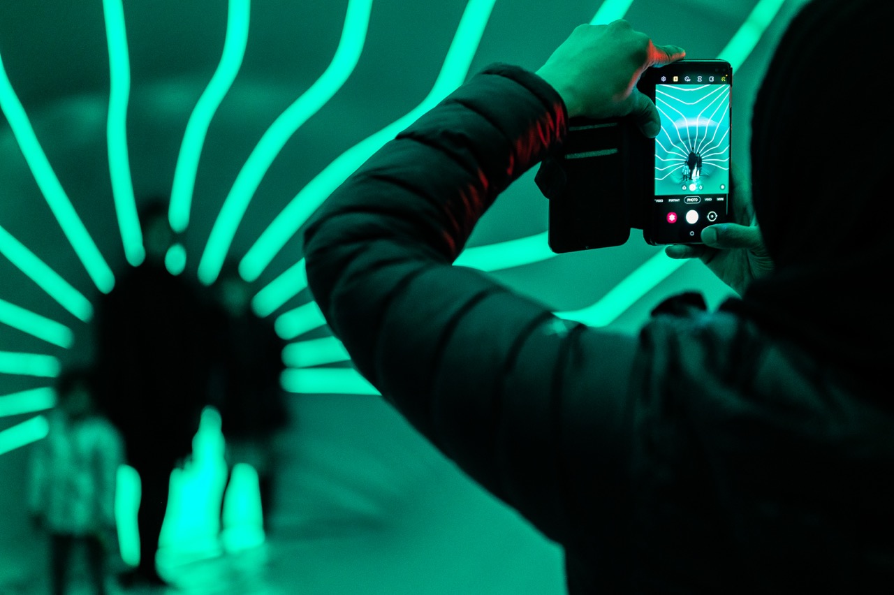 A person takes a photo with a mobile phone inside a tunnel illuminated by glowing green curved lines, with silhouetted figures walking away in the background.