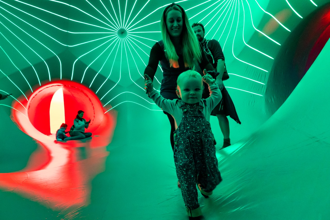 A smiling toddler walks whilst holding hands with an adult inside a large, brightly lit inflatable structure with green and red glowing patterns. Other people sit and play in the colourful background.