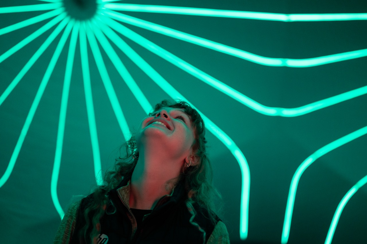 A person with curly hair looks up and smiles, illuminated by teal neon lights radiating in lines above them in a dark setting.