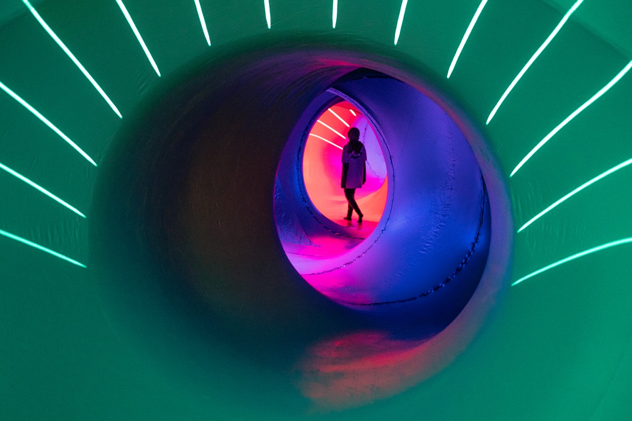A person stands inside a large, circular tunnel illuminated by vibrant neon lights in green, blue, and pink, creating a futuristic and colourful atmosphere.