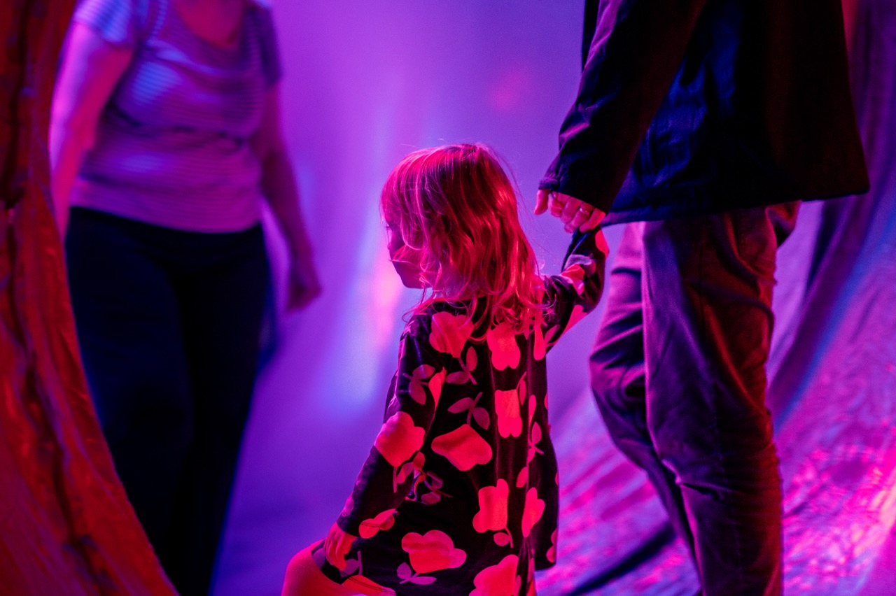 A young child in a floral dress holds an adult’s hand in a colourful, purple-lit environment, with another adult standing nearby. The scene has a dreamy, surreal atmosphere.