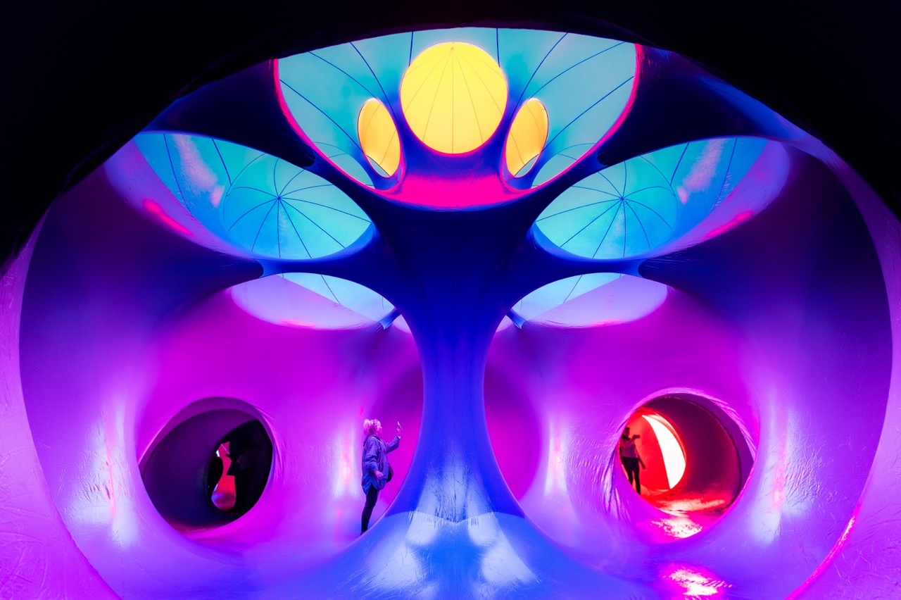 A vibrant, futuristic room with glowing pink, blue, and yellow lights features curved walls, circular openings, and two people exploring the surreal, tunnel-like space.
