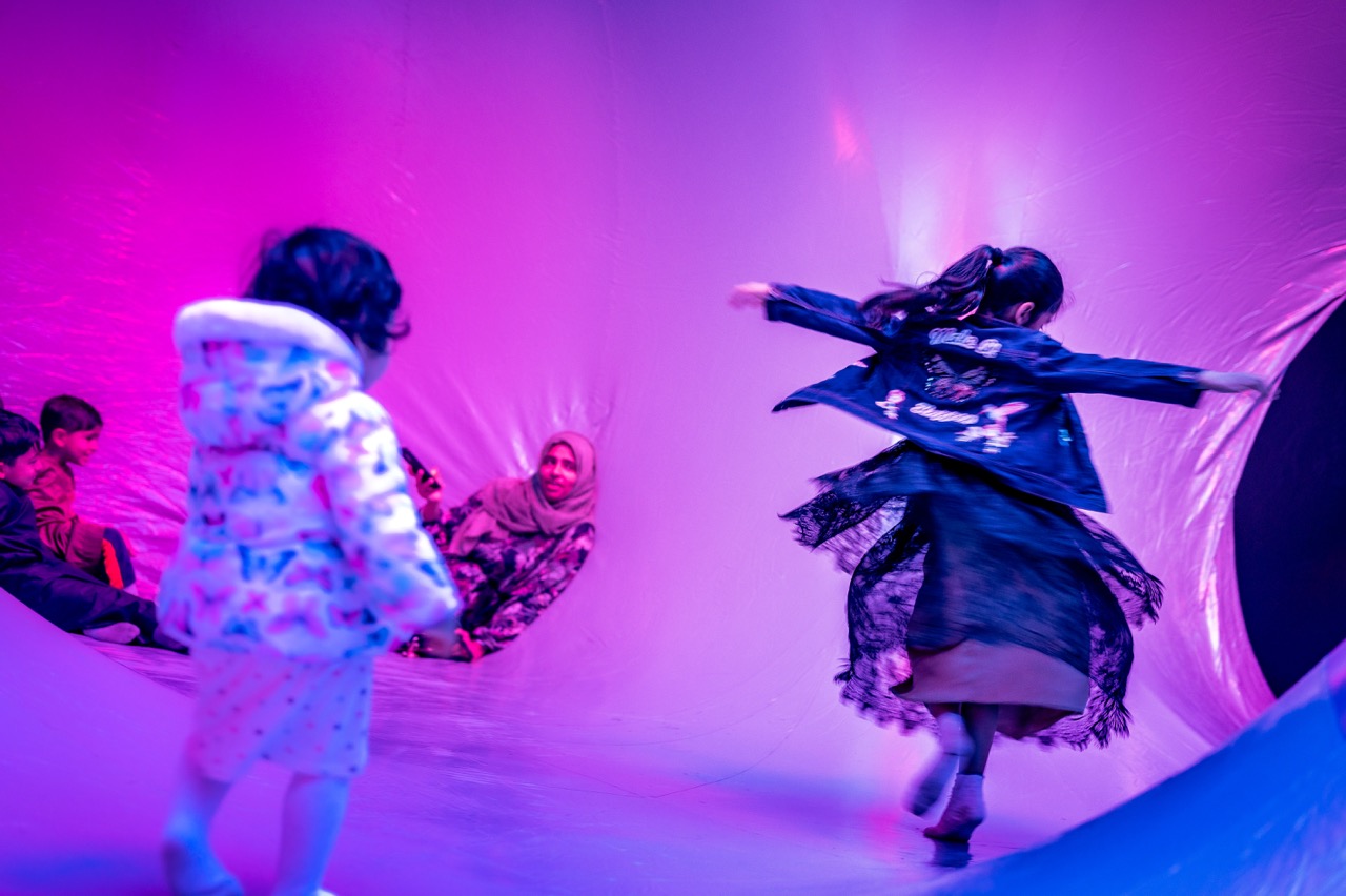 Two young girls play in a vibrant, pink and purple lit inflatable space. One girl spins with arms outstretched whilst the other stands nearby. In the background, two children and a woman sit, watching and smiling.