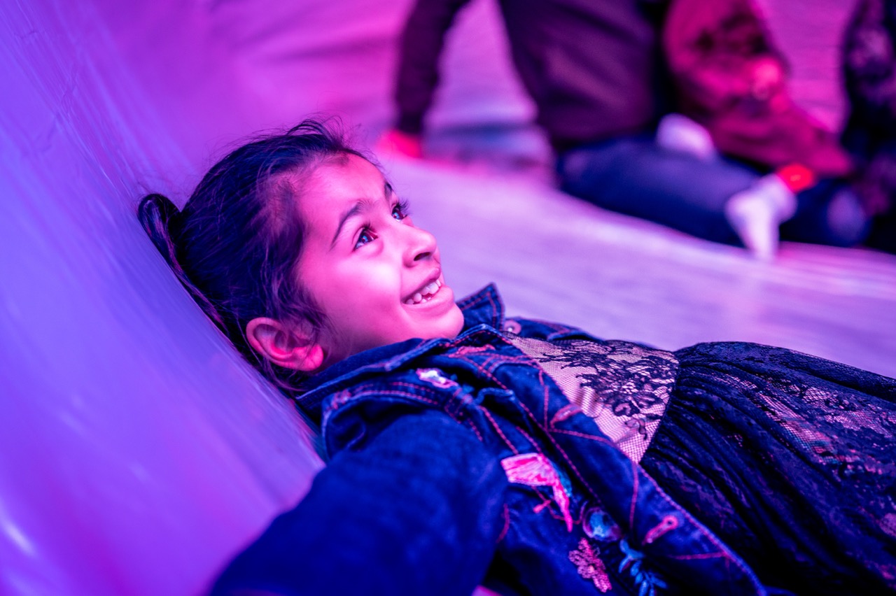 A young girl lies on her back, smiling and looking up, surrounded by purple and pink lighting. She wears a denim jacket over a dress, and other people are blurred in the background.