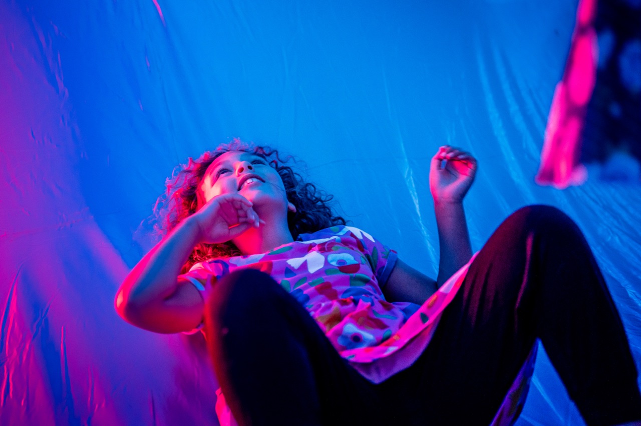 A child with curly hair lies on their back, looking upwards with a thoughtful expression. The scene is bathed in vibrant blue and pink lighting, creating a dreamy and whimsical atmosphere.