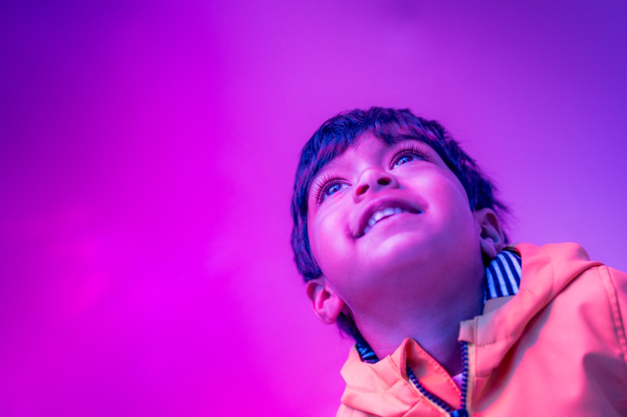 A young child in a yellow jacket looks upwards and smiles, illuminated by vibrant pink and purple lighting, against a smooth gradient background.