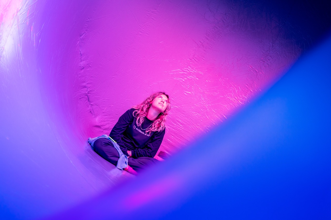 A person with long hair sits cross-legged and looks up, surrounded by curved surfaces illuminated with pink and blue light, creating an abstract, dreamlike atmosphere.