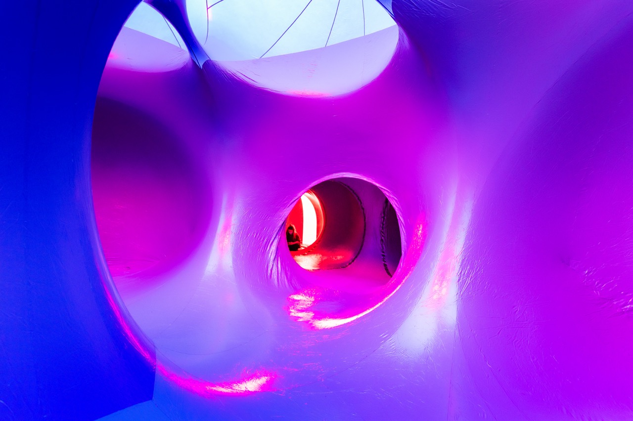 Inside a futuristic, smooth, tunnel-like structure illuminated with vibrant purple and pink lighting, several rounded openings lead to different chambers. A person is faintly visible through a circular opening in the distance.