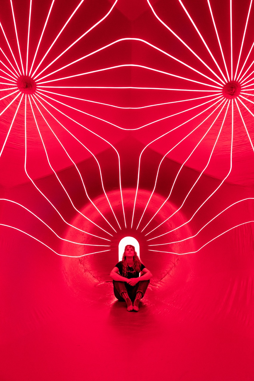 A person sits cross-legged in a tunnel illuminated by vibrant red lights and symmetrical white lines radiating from the ceiling, creating a futuristic and artistic ambience.
