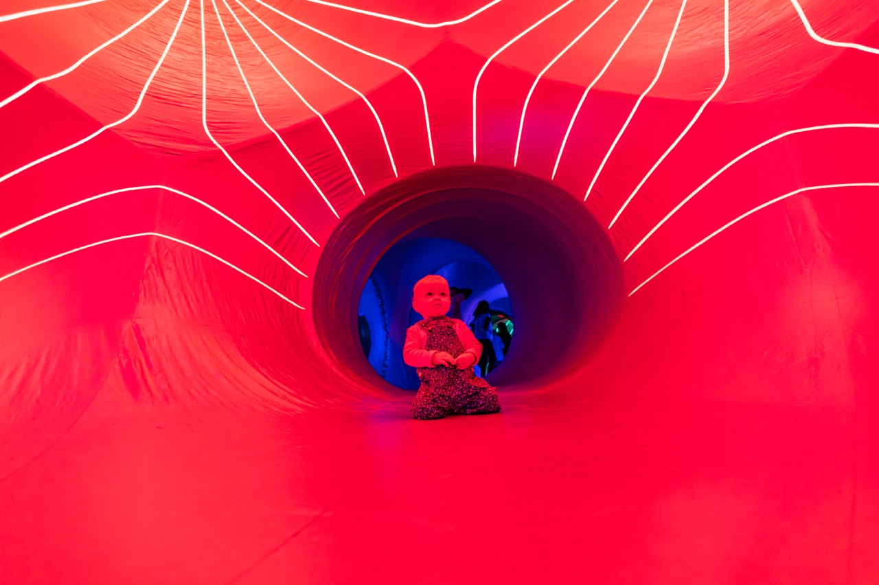 A small child sits inside a brightly lit red tunnel with glowing white lines above, creating a vibrant and playful atmosphere. Blue lighting is visible at the tunnel’s exit.
