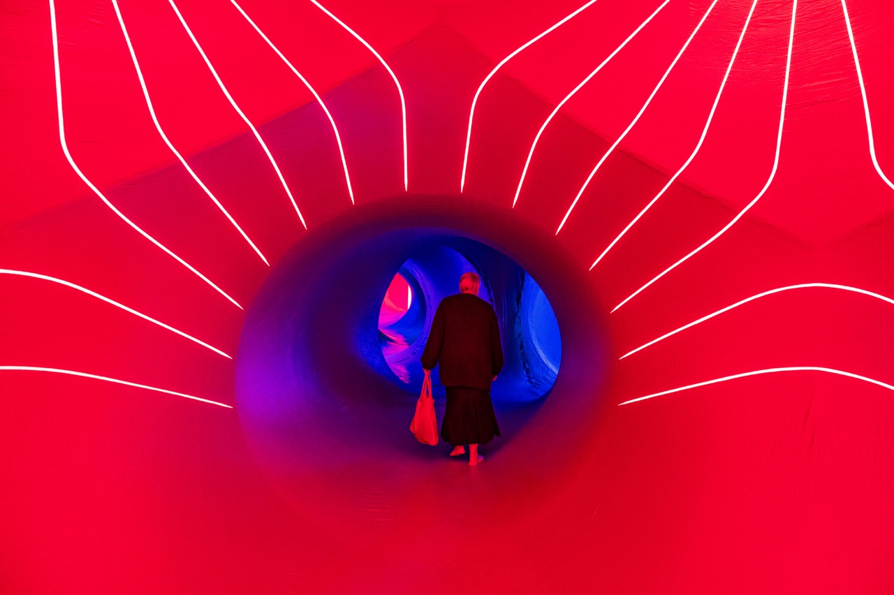 A person carrying a bag walks barefoot through a tunnel lit with vibrant red and blue lights, surrounded by curved walls with glowing white lines.