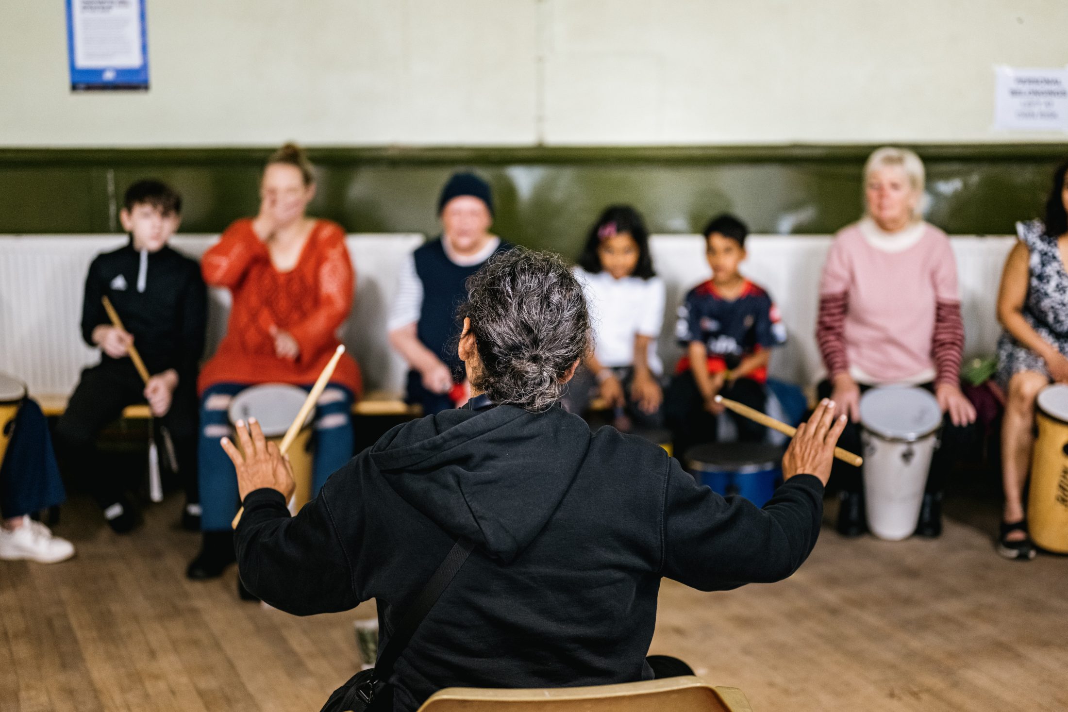 A drumming instructor seen from behind leads a group session, gesturing with drumsticks toward a diverse group of adults and children seated in a semicircle, each with a small drum, in a community hall.