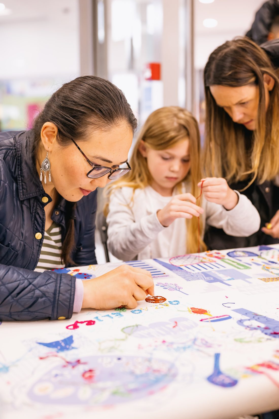Three people, including a woman and two girls, are focussed on sewing together on a large stitched cloth covered in colourful artwork. They appear to be in a bright, indoor setting.