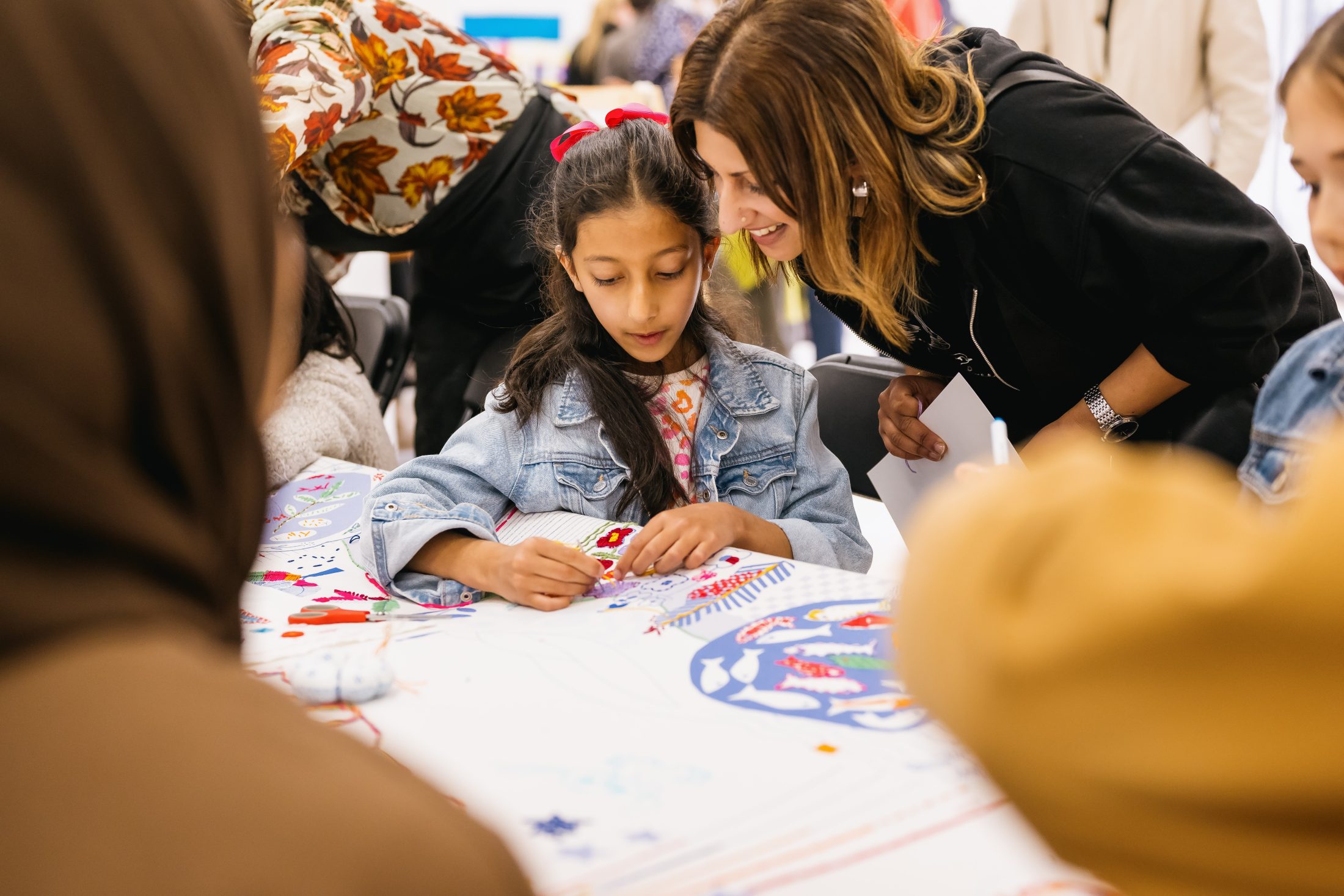 A young girl with a red bow in her hair sits at a table, creating art with colourful materials, while a smiling woman leans over to guide her. Other people are gathered round the table, participating in the activity.