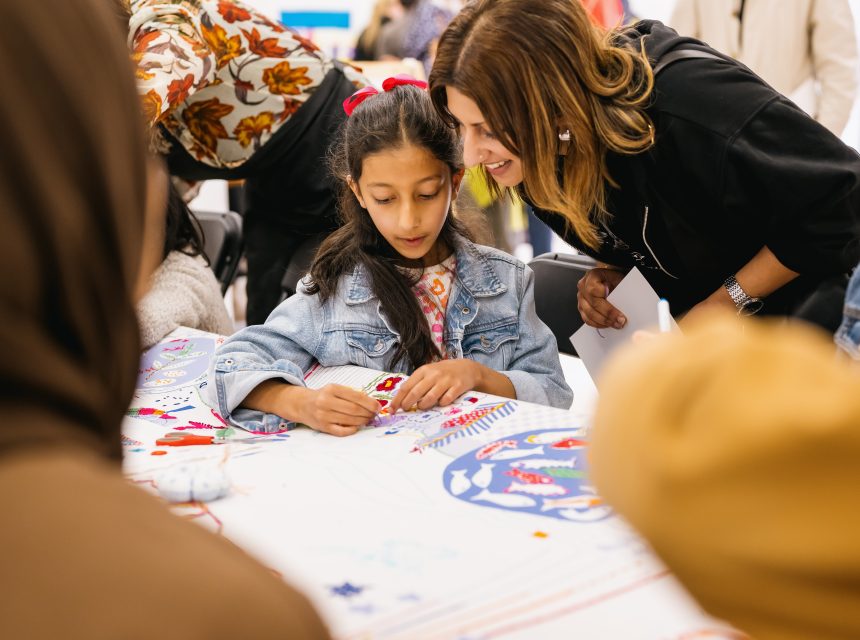A young girl with a red bow in her hair sits at a table, creating art with colourful materials, while a smiling woman leans over to guide her. Other people are gathered round the table, participating in the activity.