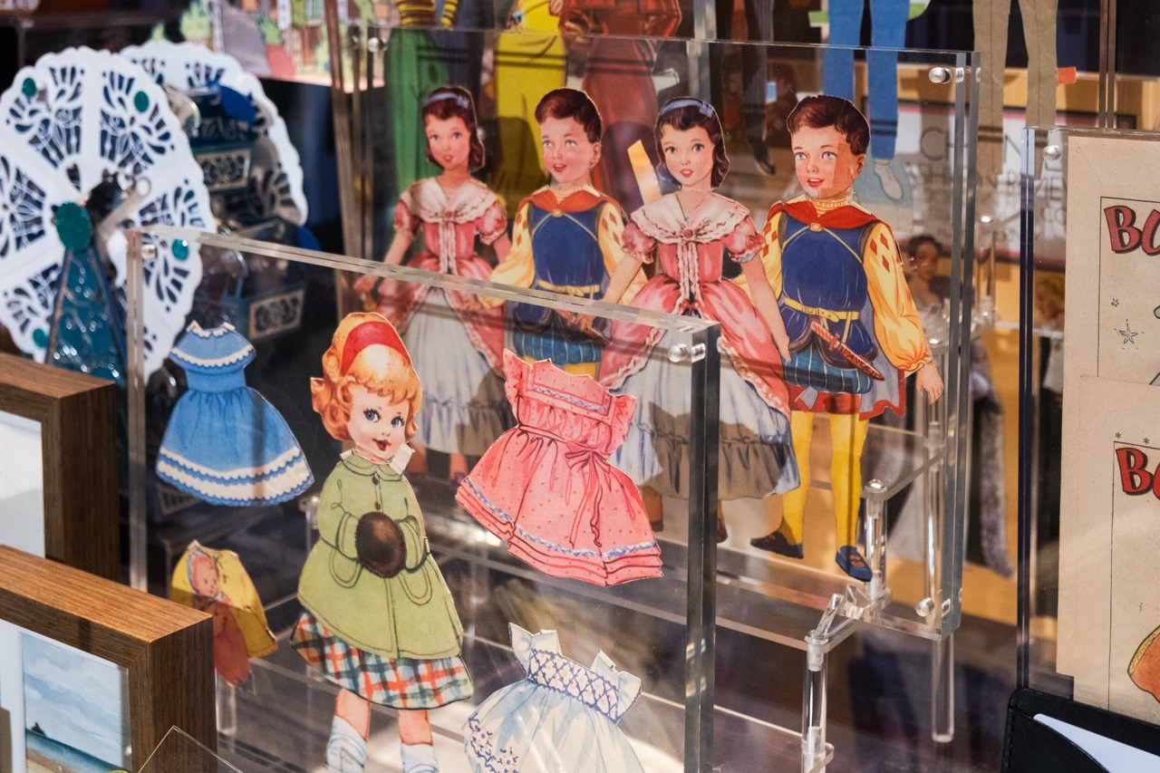 A display case filled with vintage paper dolls and colourful paper outfits, including dresses and coats, arranged neatly with some dolls standing upright and various accessories visible in the background.