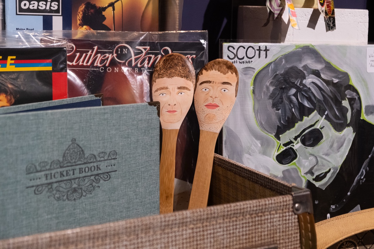 A display featuring vinyl records, a green ticket book, and two wooden spoons painted with faces. One record cover shows a painted portrait of a person in sunglasses and the word SCOTT at the top.