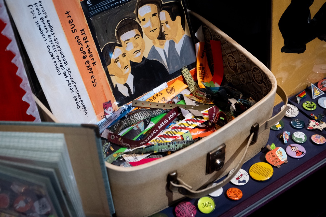 An open suitcase filled with colourful ribbons, buttons, and various memorabilia. Behind the suitcase is a painting of four people in black and white and posters with handwritten text.