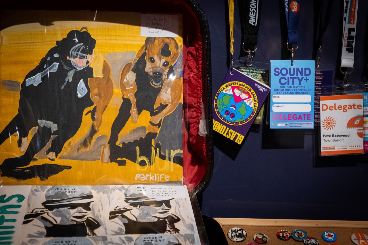 A painting of two dogs with blur parklife written below sits next to various event lanyards and badges, including ones labelled Sound City+ and Delegate, displayed against a dark background.