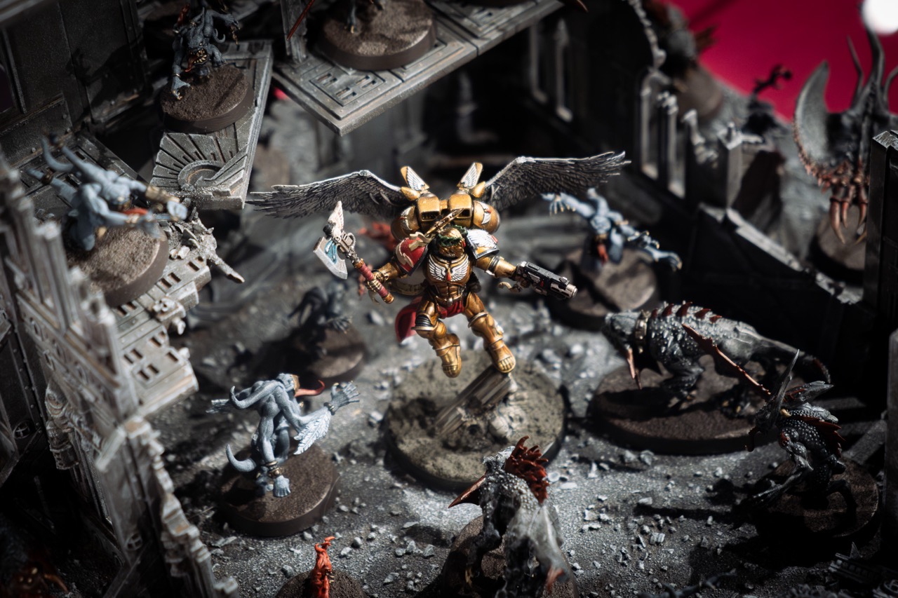 A detailed miniature diorama shows a golden-armoured, winged warrior with a jetpack and weapons battling various monstrous creatures in a ruined, gothic stone structure. Dust and debris cover the scene.