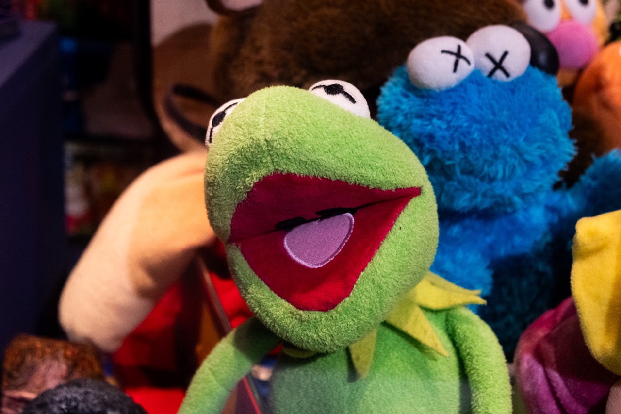 A plush toy of a green frog with an open mouth is in the foreground, with a blue monster soft toy and other colourful stuffed animals in the background.