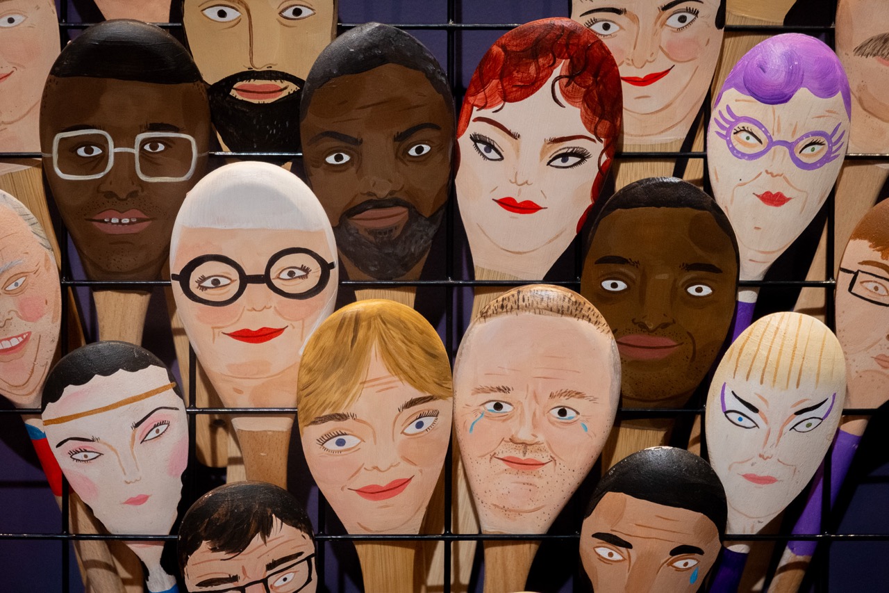 A collection of wooden spoons painted with diverse human faces, featuring different skin tones, hairstyles, and expressions, arranged upright against a metal grid background.