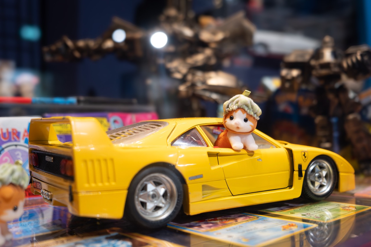 A small toy animal with a green hat sits in the driver’s seat of a yellow toy sports car, surrounded by trading cards and other toys in the background.