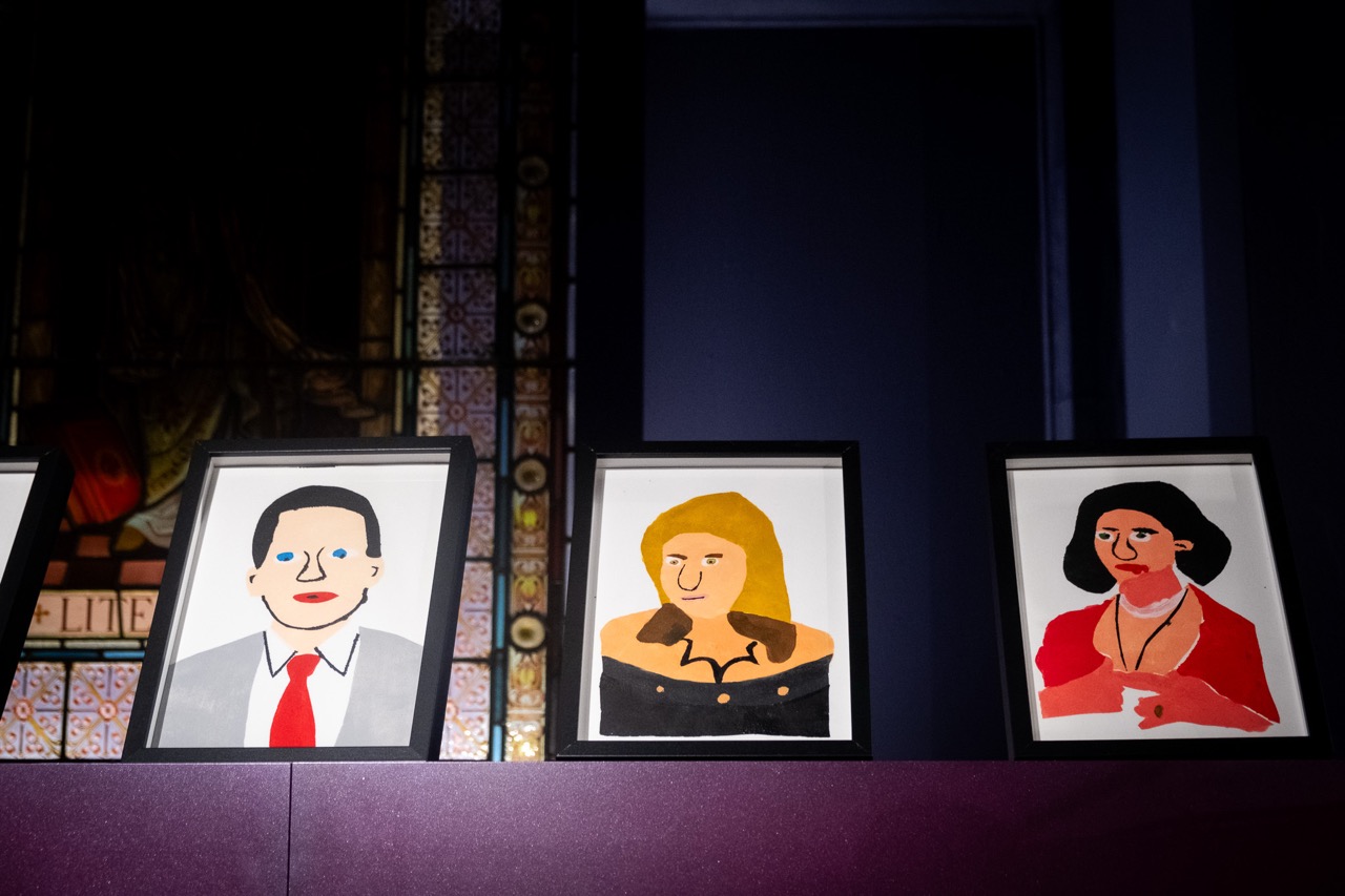 Three framed, simplistic painted portraits sit on a purple surface. Each depicts a person with distinct colours and styles against a dark background, partially illuminated by light. A patterned window is visible to the left.