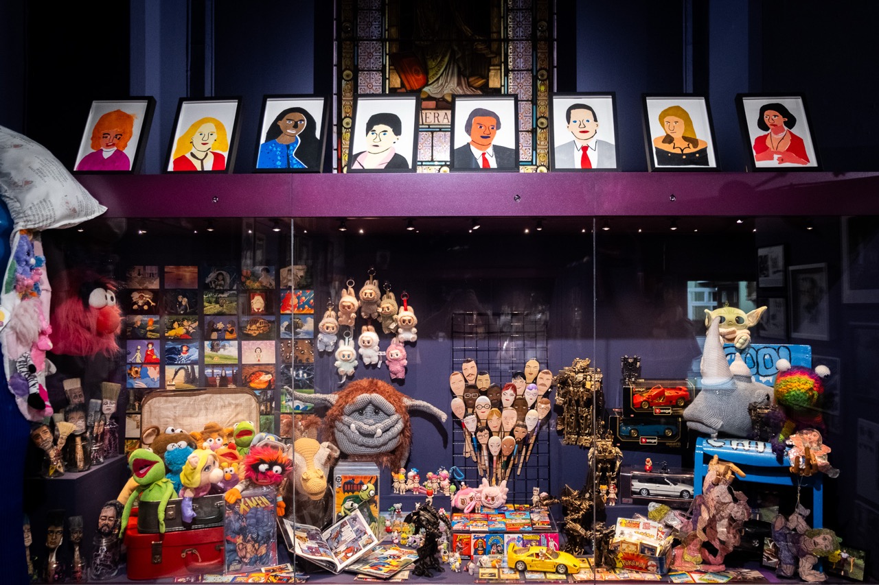 A display case filled with colourful puppets, toys, and memorabilia, with framed cartoon portraits of people lined up on the shelf above. A stained glass window is partially visible behind the portraits.