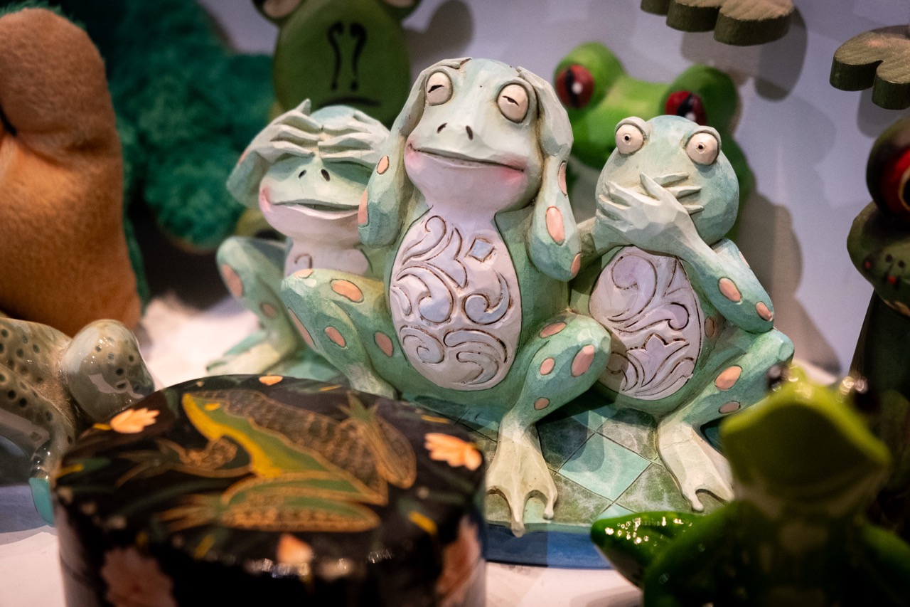 Three decorative frog figurines display see no evil, hear no evil, speak no evil gestures, surrounded by other frog-themed items, including a box with a frog illustration and stuffed frogs in the background.